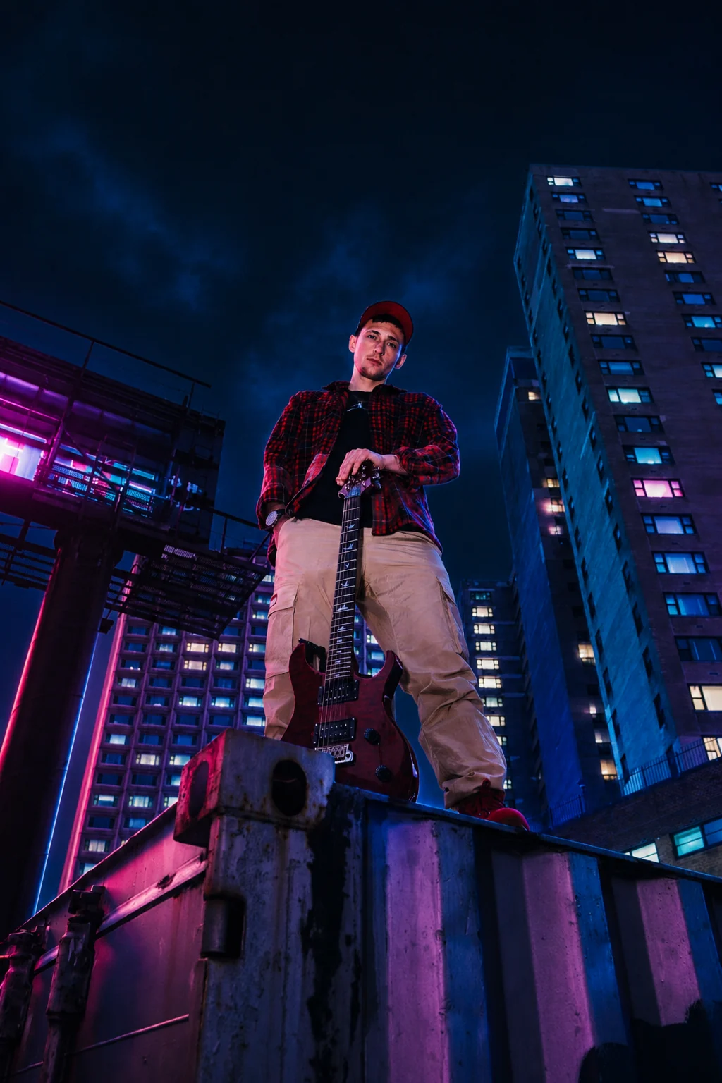 Kaden Quinn holding an electric guitar on a rooftop at night, neon skyscraper lights behind him